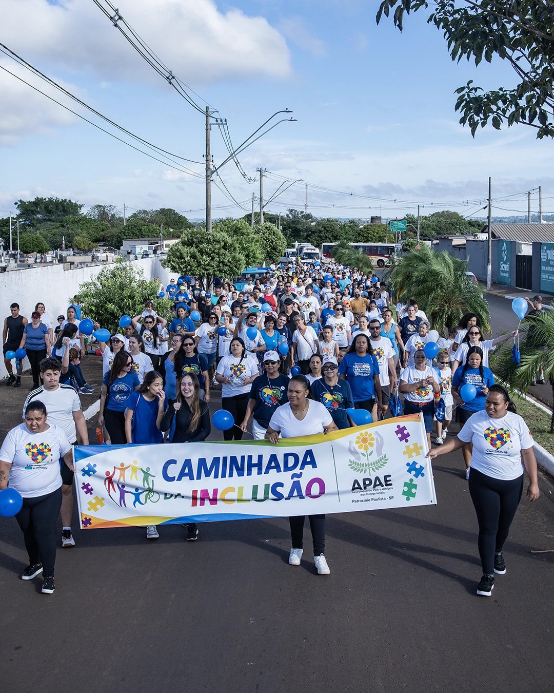 Multidão de camiseta branca e azul caminhando atrás da faixa da Caminhada da Inclusão da APAE