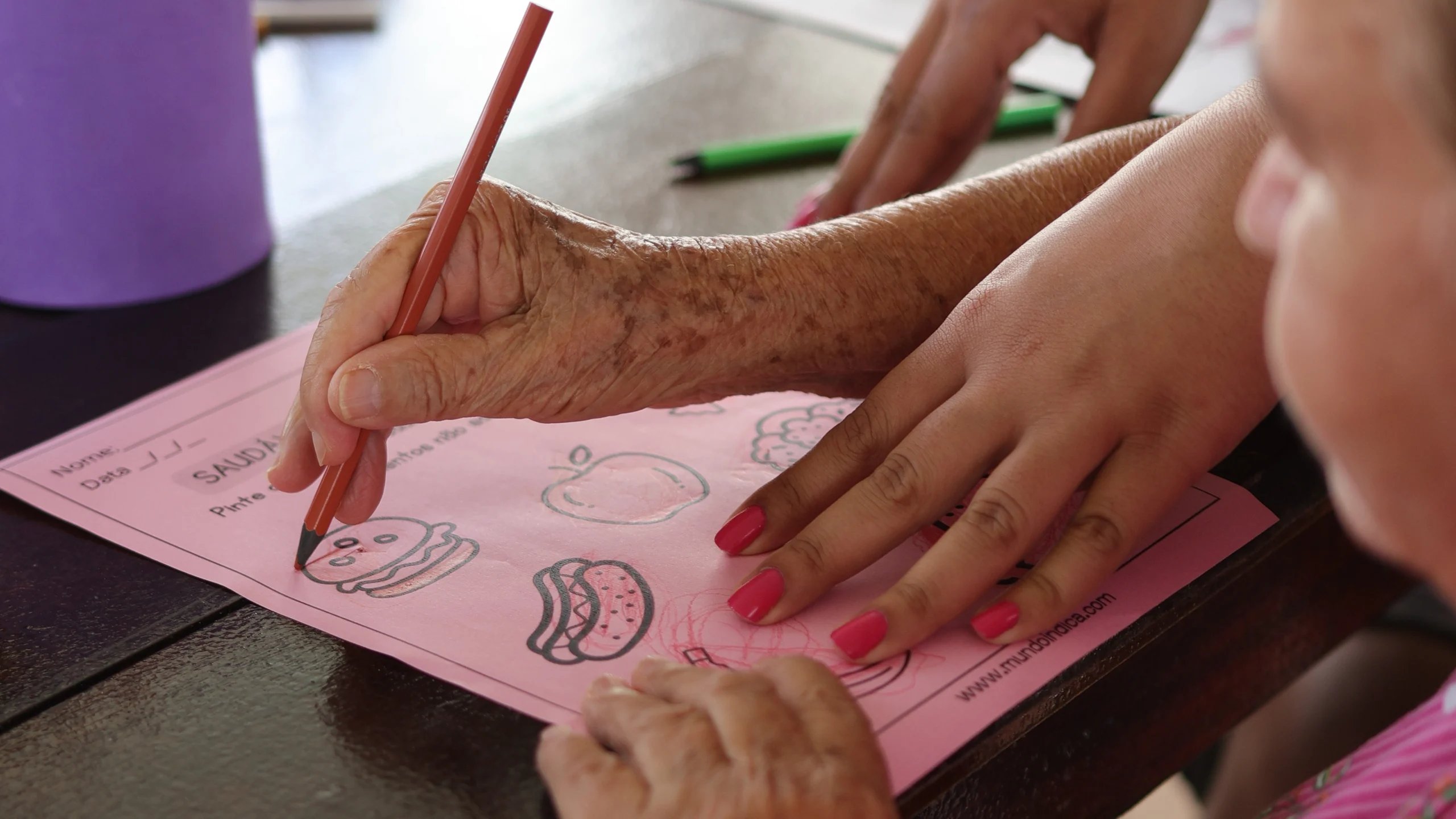 Mãos de uma atendida do Centro-Dia desenhando, acompanhada por uma educadora