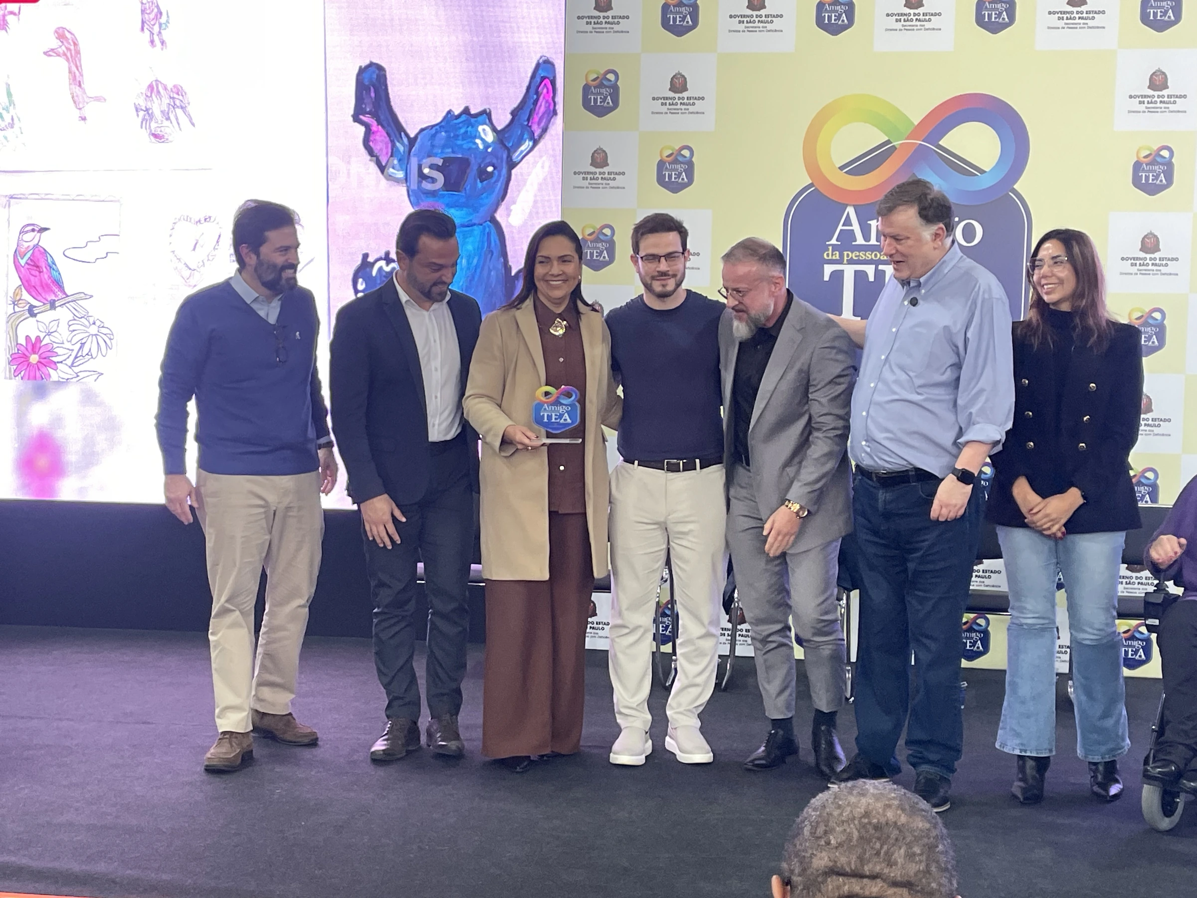Representante da APAE no palco, ao lado de autoridades, segurando o Selo Amigo da Pessoa com TEA
